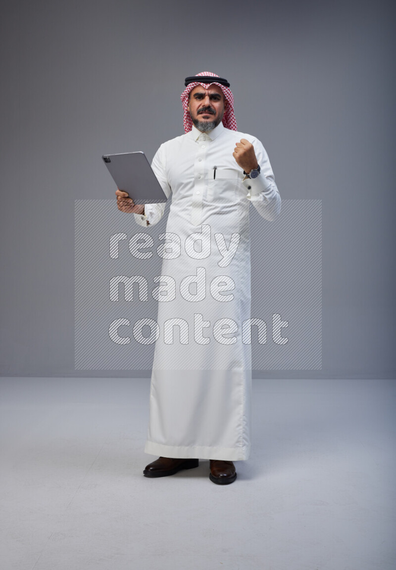 Saudi man Wearing Thob and red Shomag standing working on tablet on Gray background