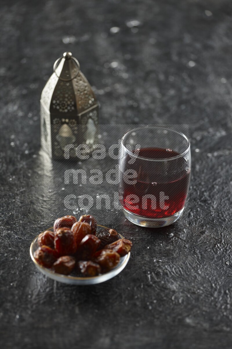 A silver lantern with different drinks, dates, nuts, prayer beads and quran on textured black background