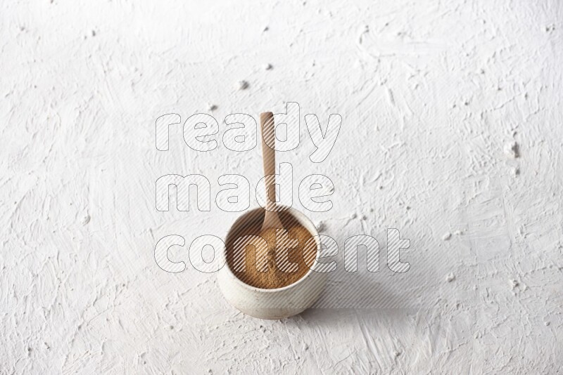 Ceramic beige bowl full of cinnamon powder with a wooden spoon on a textured white background