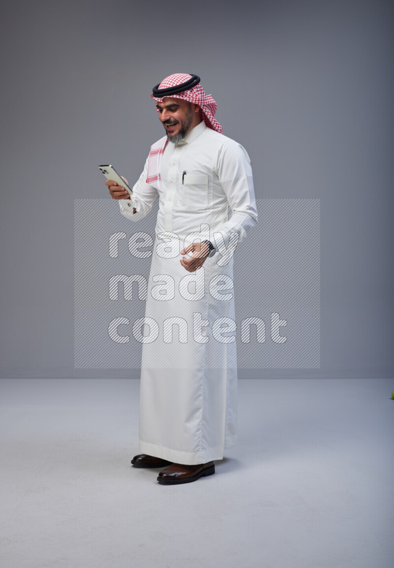 Saudi man Wearing Thob and red Shomag standing texting on phone on Gray background