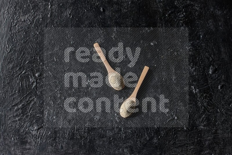 2 wooden spoons full of white pepper powder on textured black flooring