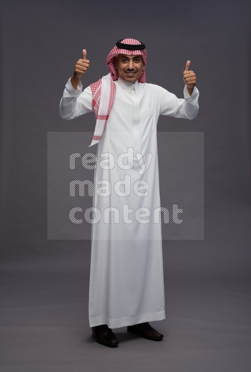 Saudi man wearing thob and shomag standing interacting with the camera on gray background