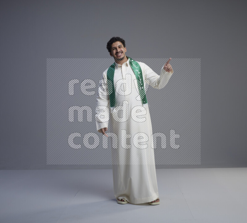 A Saudi man standing wearing thob and Saudi flag scarf on gray background