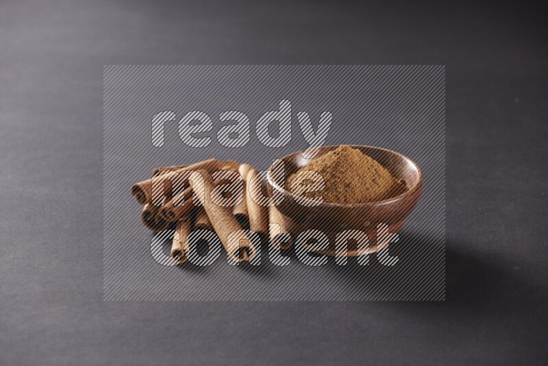Cinnamon sticks stacked beside a wooden bowl full of cinnamon powder on black background