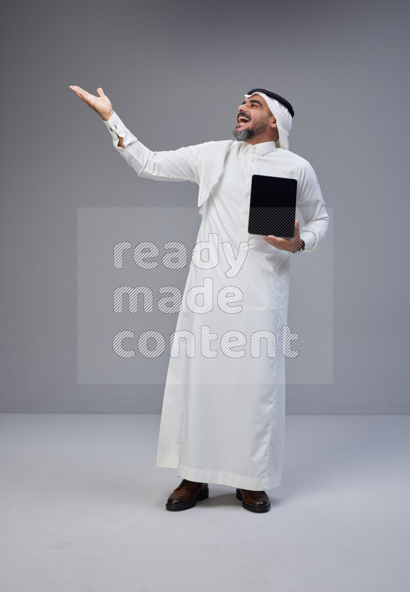 Saudi man Wearing Thob and white Shomag standing showing tablet to camera on Gray background