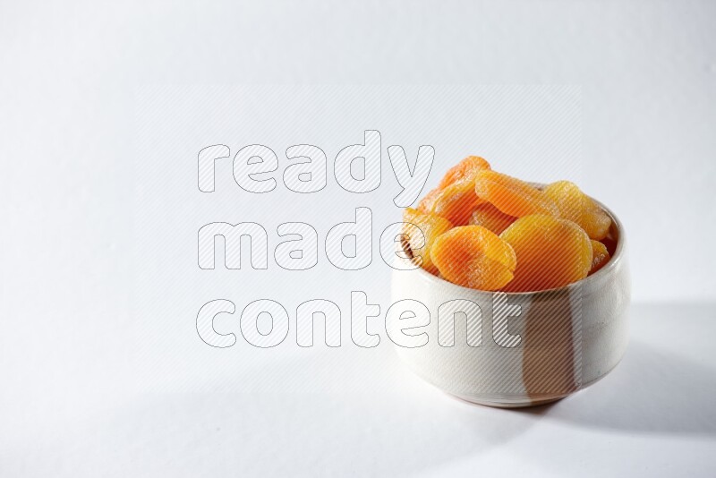 A beige ceramic bowl full of dried apricots on a white background in different angles