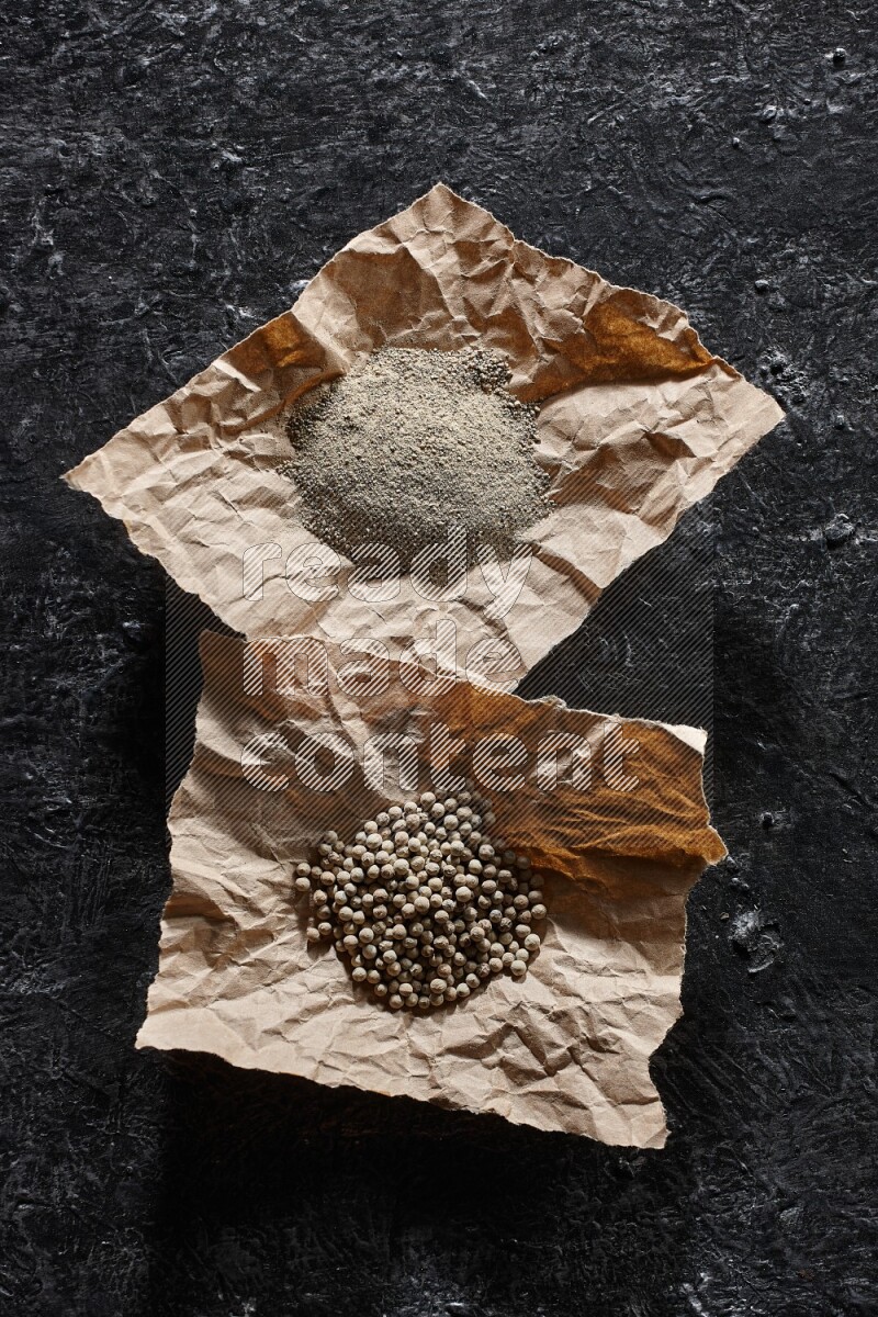 White pepper beads and powder in 2 crumpled paper on textured black flooring