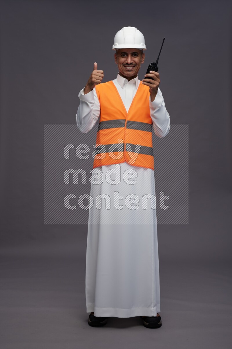 Saudi man wearing thob with engineer vest standing holding walkie-talkie on gray background