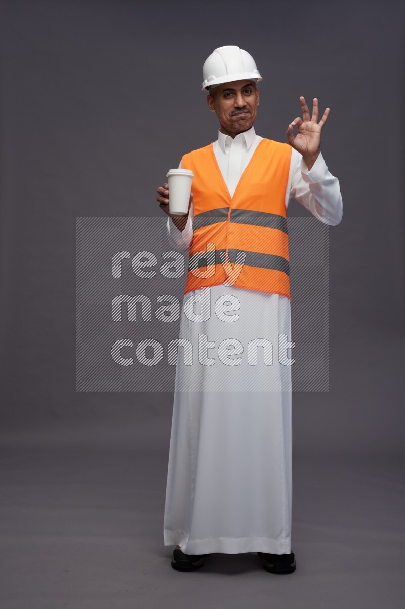 Saudi man wearing thob with engineer vest standing holding paper cup on gray background