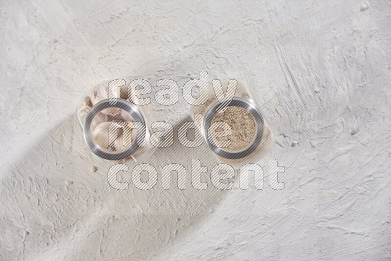 2 glass spice jars one full of garlic powder and the other full of cloves on a textured white flooring