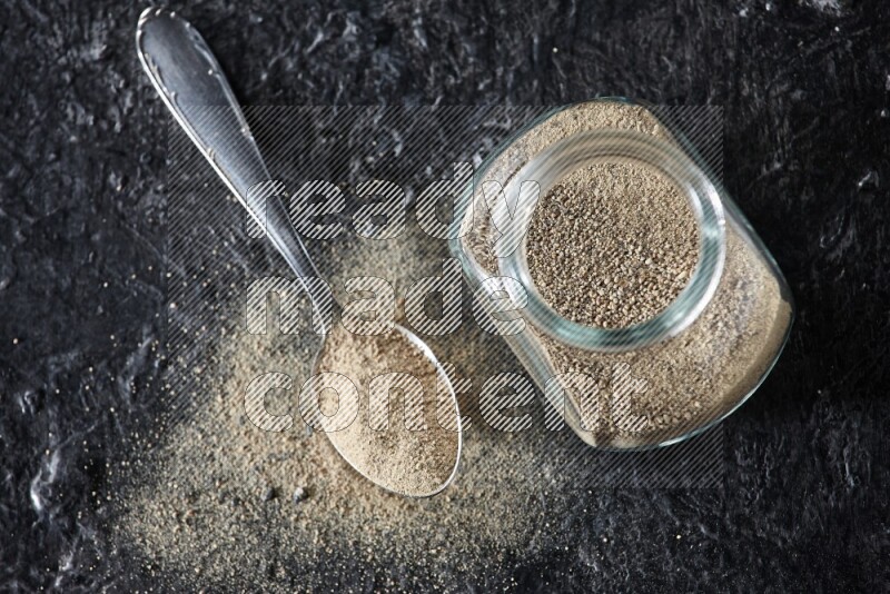 Herbal glass jar and metal spoon full of white pepper powder on textured black flooring