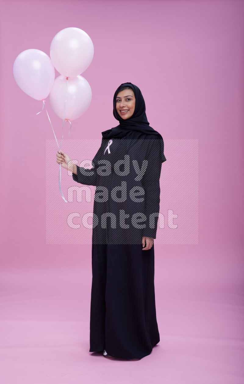 Saudi woman wearing pink ribbon on Abaya standing holding balloons on plain pink background