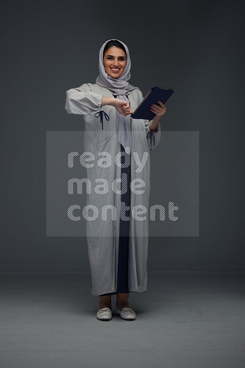A Saudi woman wearing a light gray Abaya and head scarf standing holding a phone and pointing with the other hand eye level on a grey background