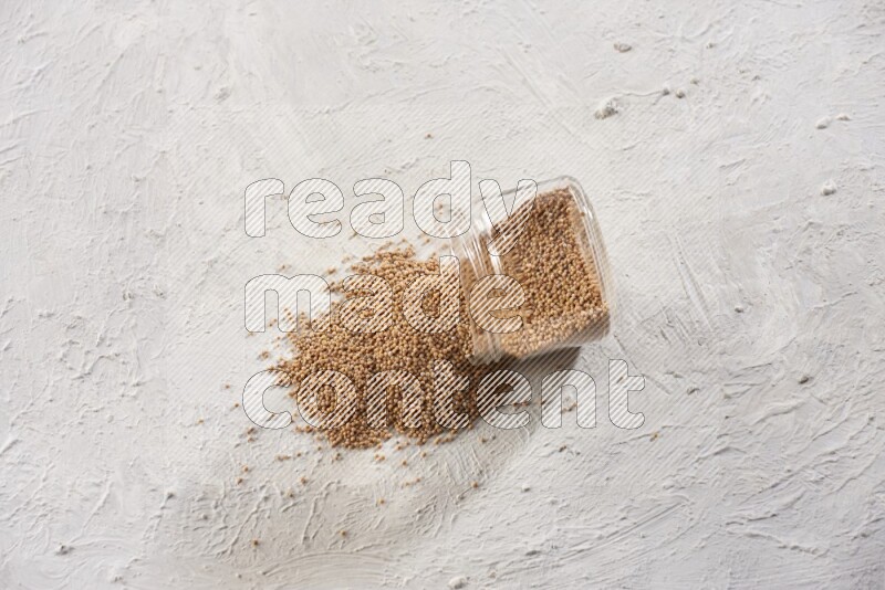 A glass jar full of mustard seeds and jar is flipped with seeds spread out on a textured white flooring