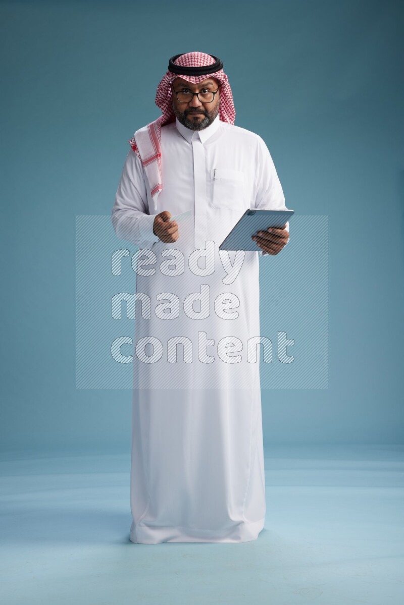 Saudi Man with shimag Standing holding ATM card while working on tablet on blue background