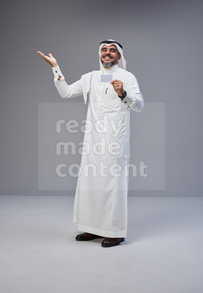 Saudi man Wearing Thob and white Shomag standing holding ATM card on Gray background
