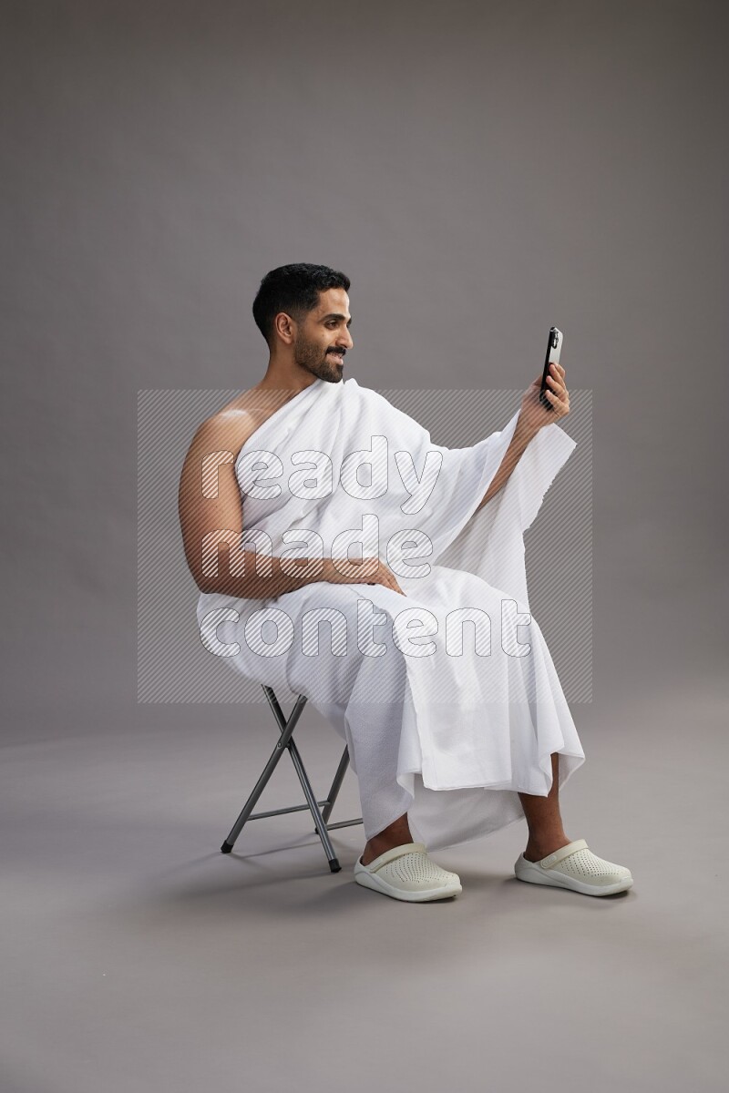 A man wearing Ehram sitting on chair taking selfie on gray background