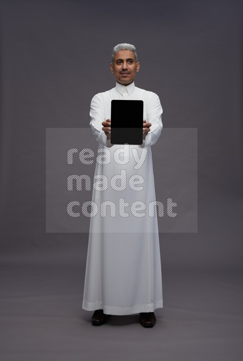 Saudi man wearing thob standing showing tablet to camera on gray background