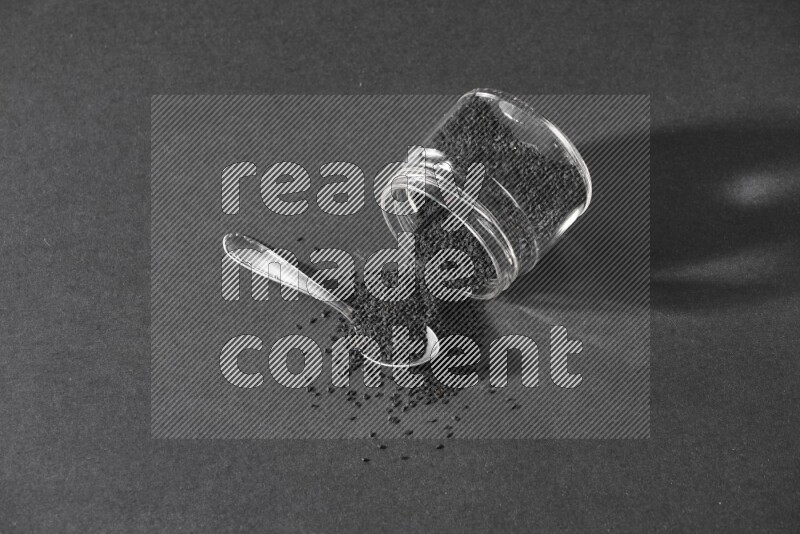 وعاء زجاجي مقلوب ممتلئ ببذور حبة البركة والبذور متناثرة بجانبه مع ملعقة معدنية ممتلئة بالبذور على أرضية سوداء