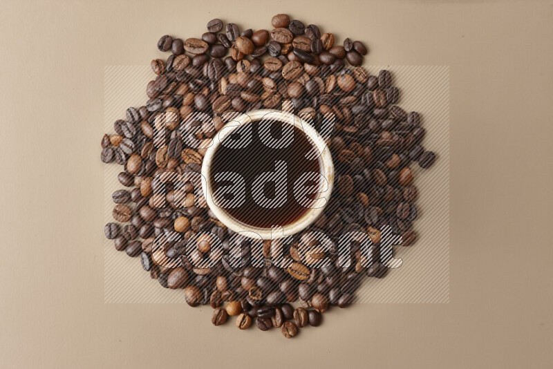 A beige pottery cup of coffee surrounded by roasted coffee beans on beige background