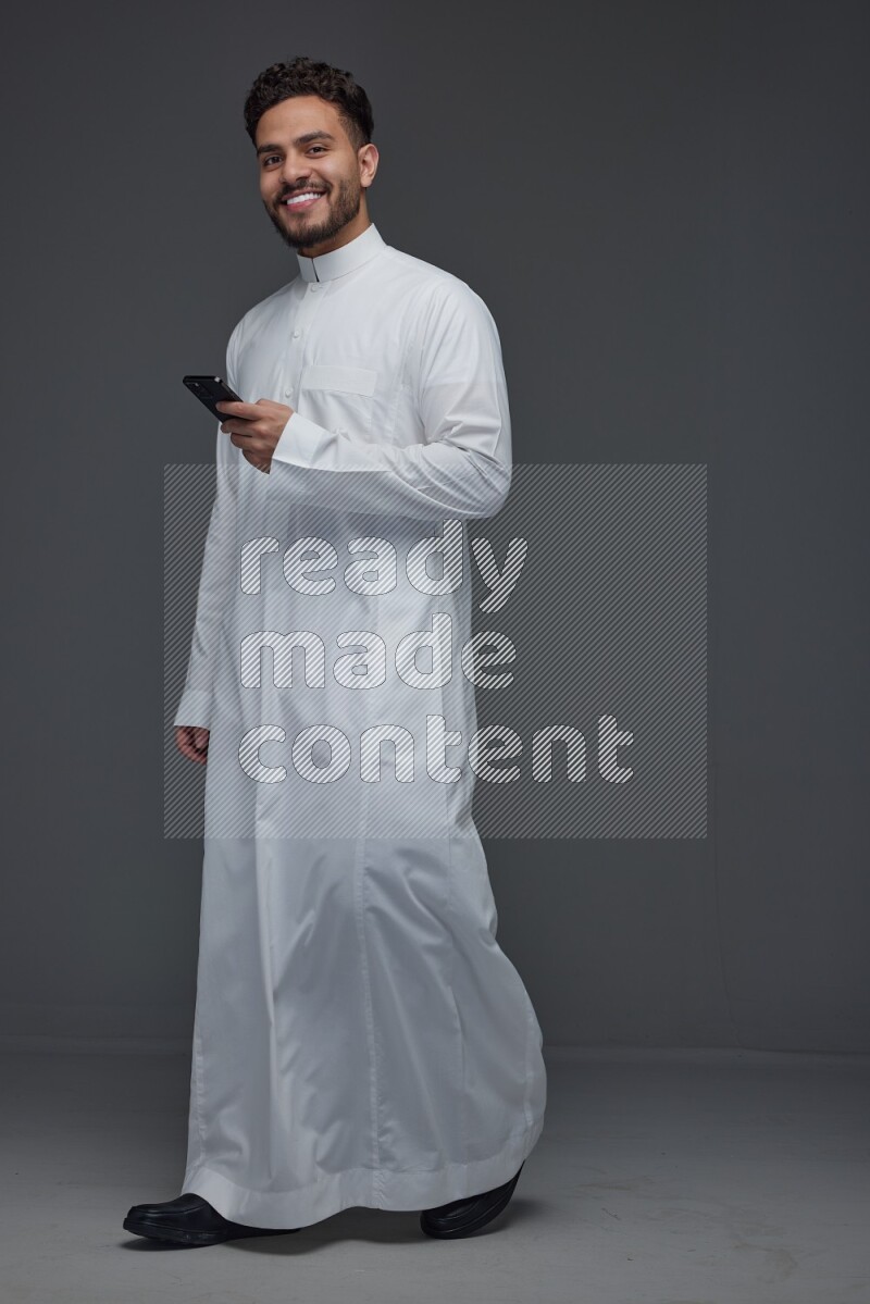 A Saudi man wearing Thobe standing and using his phone eye level on a gray background