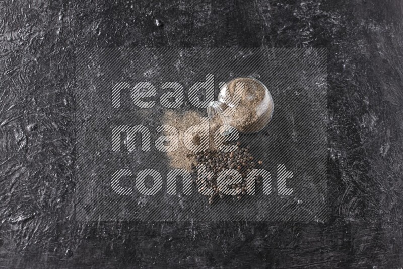 A flipped glass spice jar full of black pepper powder with pepper beads on textured black flooring