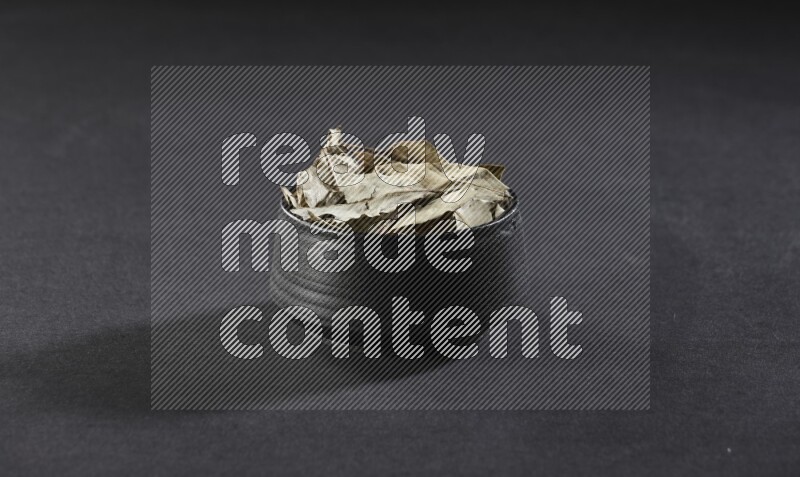 A black pottery bowl full of dried bay leaves on black flooring