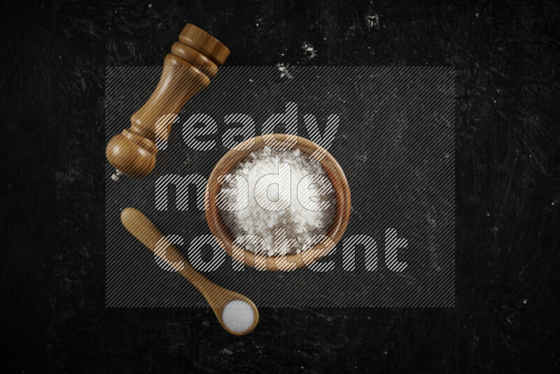 A wooden bowl and spoon filled with white sea salt and wooden grinder beside them on black background