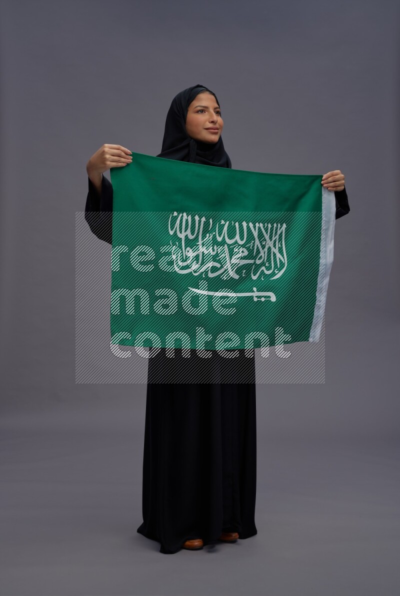 Saudi woman wearing Abaya standing holding Saudi flag on gray background