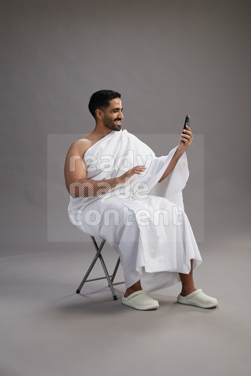 A man wearing Ehram sitting on chair taking selfie on gray background