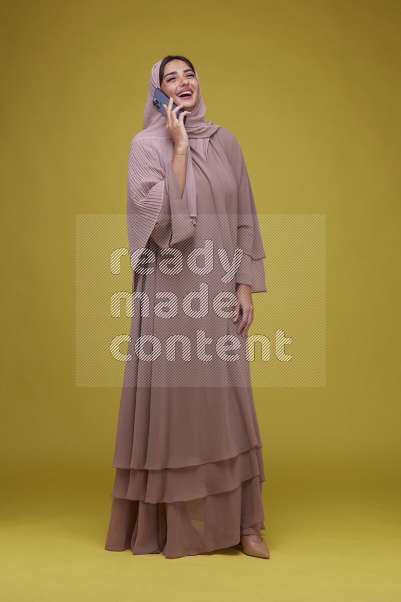 A woman having a call on a Yellow Background wearing Brown Abaya with Hijab