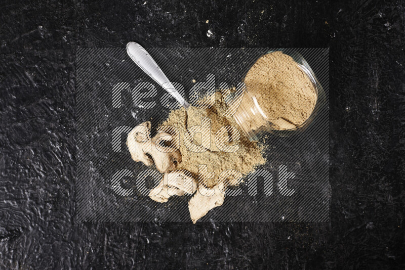 A glass jar full of ground ginger powder flipped with some spilling powder on black background