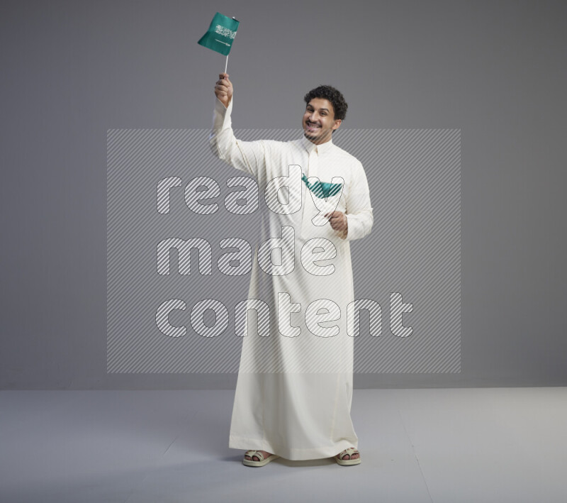 A Saudi man standing wearing thob holding small Saudi flag on gray background