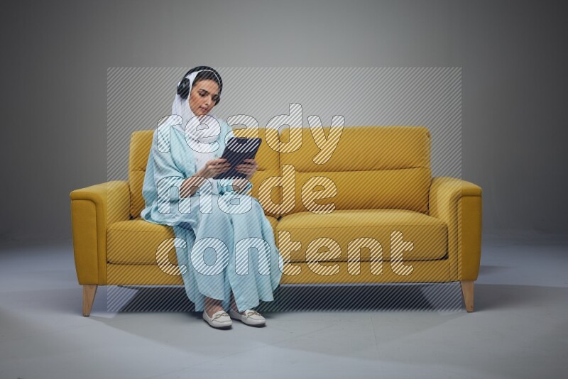 A Saudi woman wearing a light blue Abaya and a white head scarf sitting on a yellow sofa and holding an iPad while wearing headphone eye level on a grey background