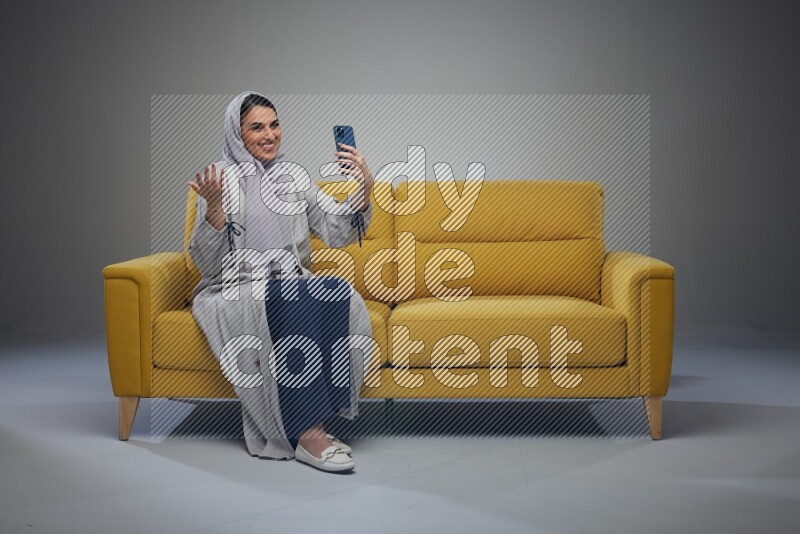 A Saudi woman wearing a light gray Abaya and head scarf sitting on a yellow sofa and making a video call eye level on a grey background