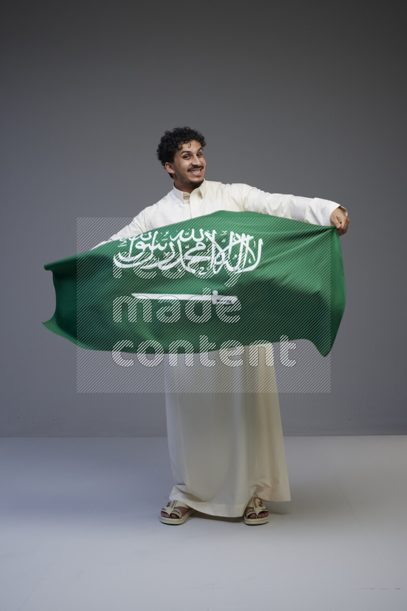 A Saudi man standing wearing thob holding big Saudi flag on gray background