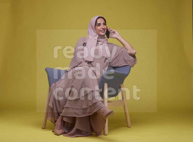 A woman having a call on a blue Chair on a Yellow Background wearing Brown Abaya with Hijab