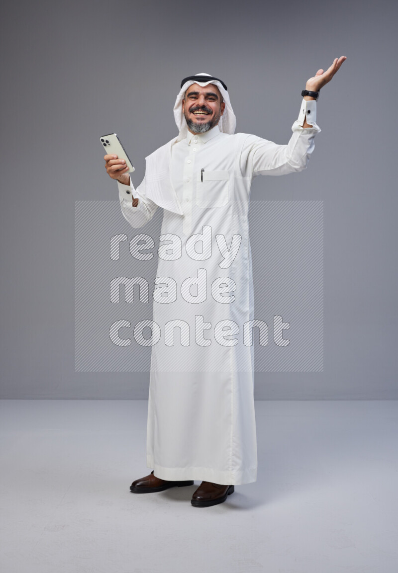 Saudi man Wearing Thob and white Shomag standing texting on phone on Gray background