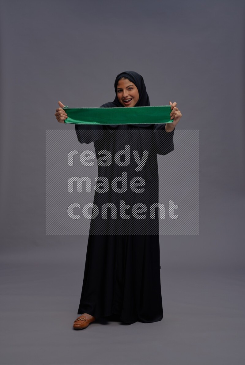 Saudi woman wearing Abaya standing holding Saudi flag on gray background