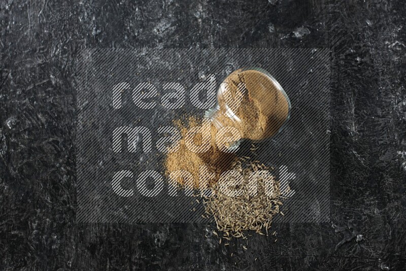 وعاء زجاجي مقلوب ممتلئ ببودرة الكمون مع بودرة متناثرة وحبوب الكمون على خلفية سوداء