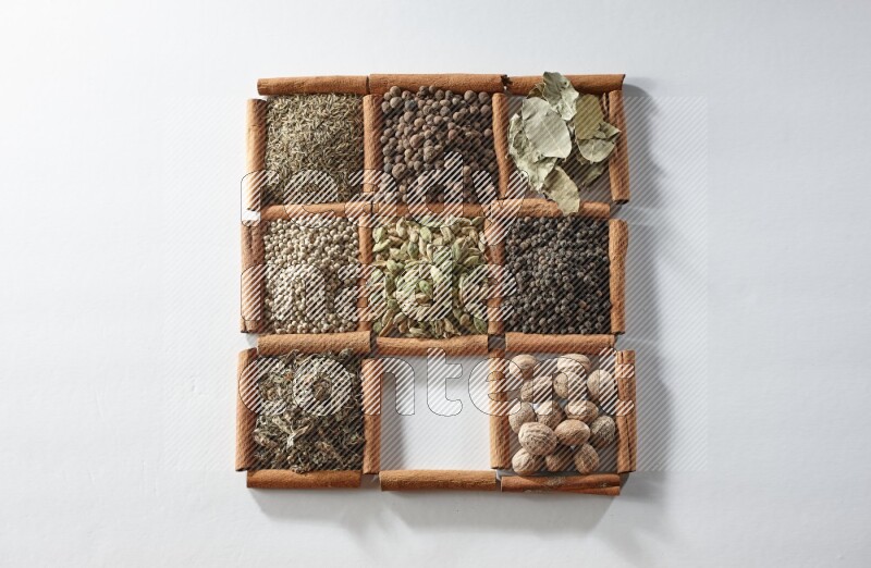 9 مربعات من أعواد القرفة ممتلئة بالهيل في المنتصف محاطة بجوزة الطيب والملح والريحان المجفف والفلفل الأبيض والكمون وبهار الفلفل الحلو وأوراق الغار والفلفل الأسود على أرضية بيضاء