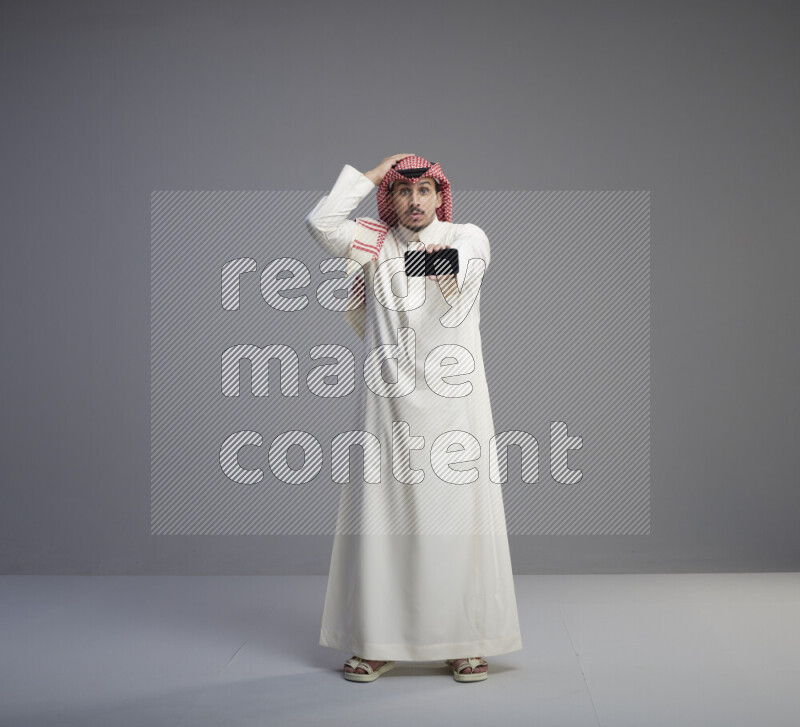 A Saudi man standing wearing thob and red shomag showing phone to camera on gray background