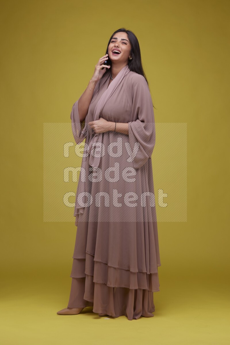 A woman Calling on a Yellow Background wearing Brown Abaya