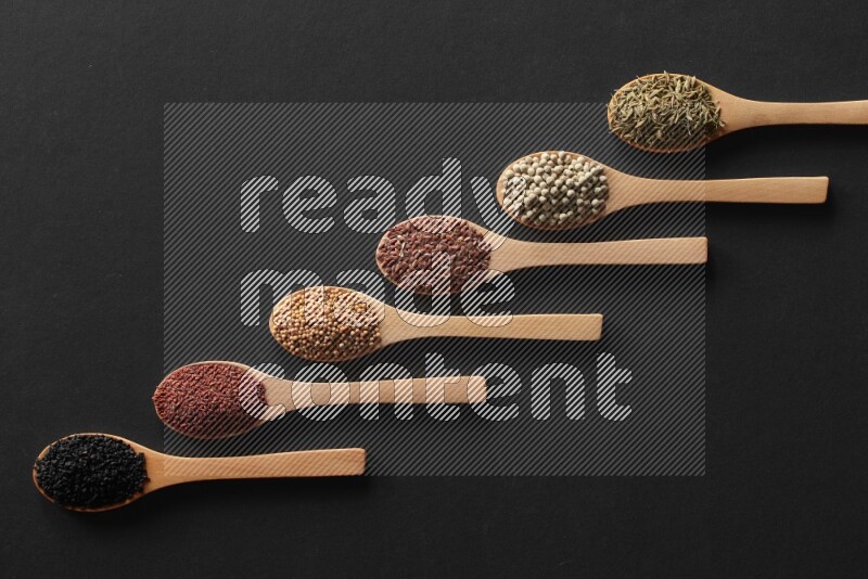 wooden spoons filled with white peppers, mustard seeds, black seeds, garden cress, cumin and flax on black flooring