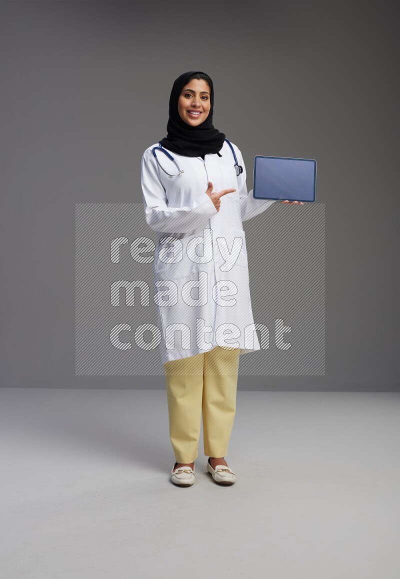 Saudi woman wearing lab coat with stethoscope standing showing tablet to camera with sign in the back on Gray background