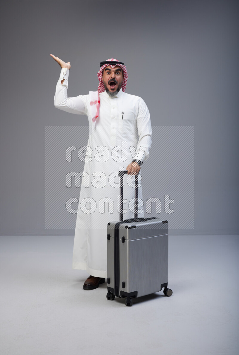 Saudi man wearing Thob and red Shomag standing holding Travel bag on Gray background