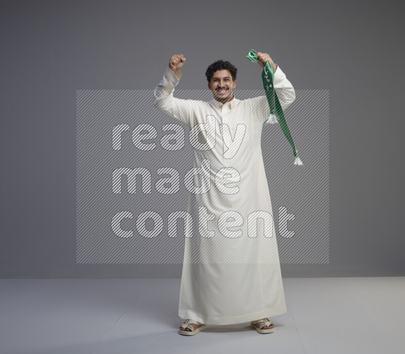 A Saudi man standing wearing thob holding Saudi flag scarf on gray background