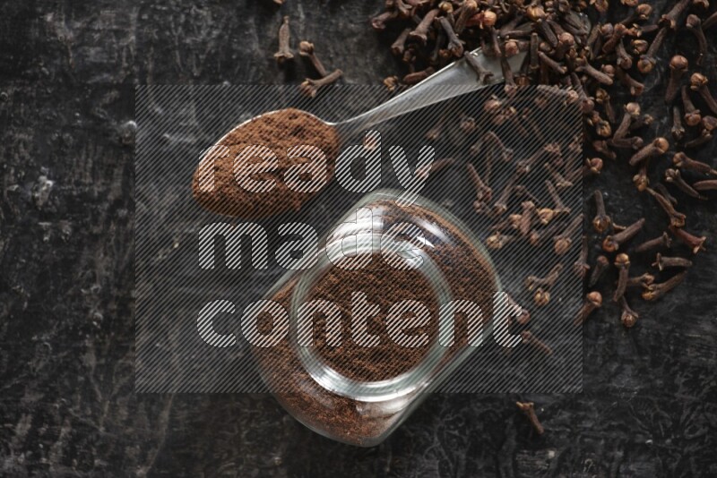 A glass spice jar and a metal spoon full of cloves powder with cloves spread on textured black flooring