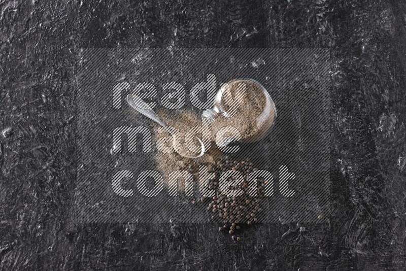 A flipped glass spice jar full of black pepper powder with a metal spoon full of powder and pepper beads on textured black flooring