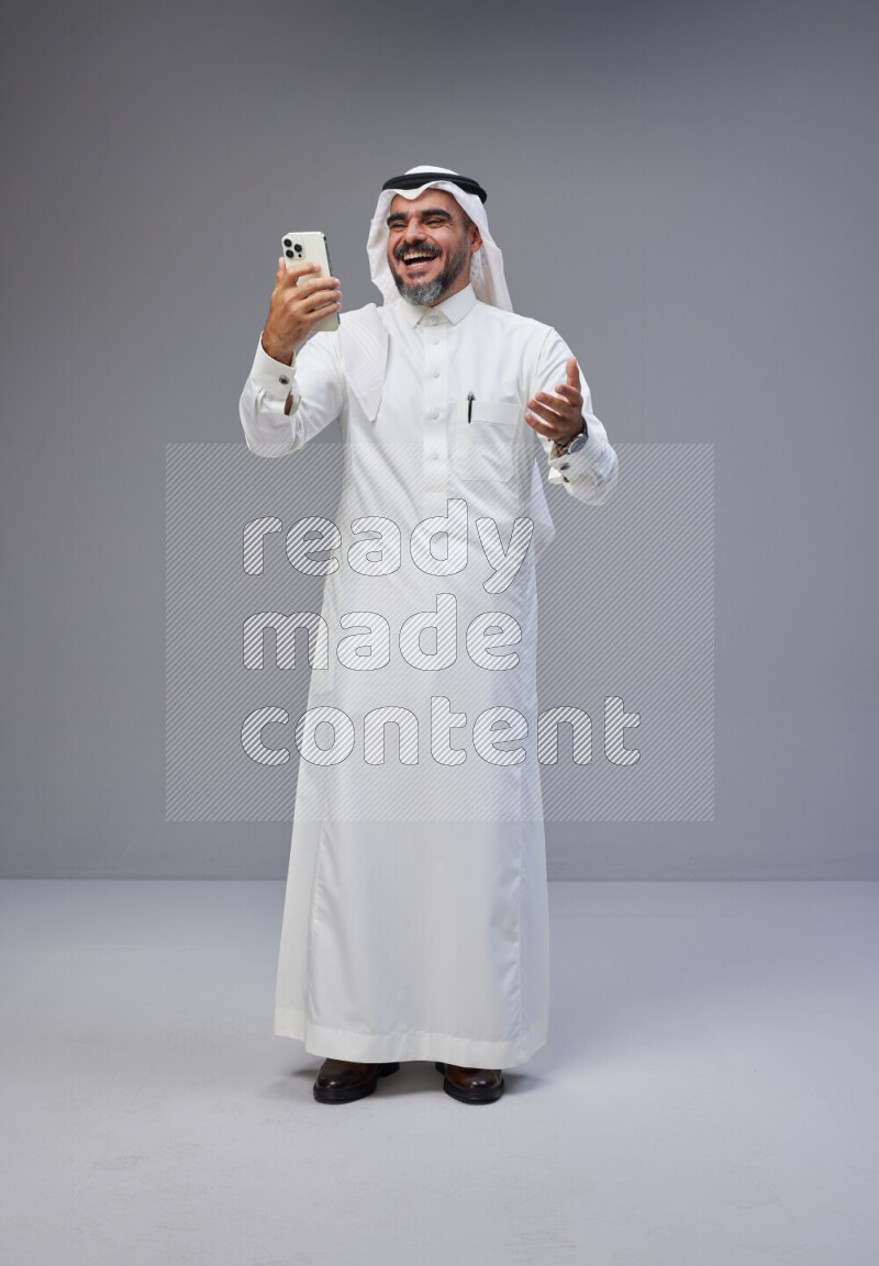 Saudi man Wearing Thob and white Shomag standing texting on phone on Gray background